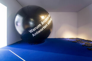 Ein großer, schwarzer Ball mit der weißen Aufschrift „Warum ändern sich Antworten ständig?“ liegt in einem Raum mit weißen Wänden und blauem Boden. Viele kleine schwarze Kugeln sind auf dem Boden verstreut. Licht fällt durch ein Fenster links und erzeugt Schatten.